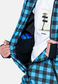 Chaqueta exterior a cuadros azules y negros desabrochada, mostrando una camiseta blanca con un estampado gráfico y un par de gafas de protección guardadas en el bolsillo.