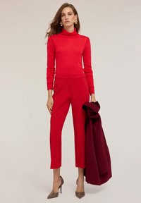Pulover roșu cu guler înalt, pantaloni roșii adaptați cu tiv scurt, palton maron închis și pantofi cu toc înalt bej. Textură netedă și design ajustat.