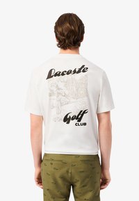 Camiseta blanca de algodón con un diseño gráfico en la parte de atrás que presenta el texto "Lacoste Golf Club" y una ilustración de un golfista en un entorno de parque.