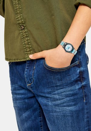 Grünes Baumwollhemd mit Knopfdetails, kombiniert mit blauen Jeans. Trägt eine Uhr mit weißem Zifferblatt und hellblauem Armband mit Mustern.