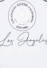 Bela tkanina s okroglo oznako, ki vsebuje napis "GUESS CALIFORNIA" in "EST. 1981", poudarjena z črnimi in metalno zlatimi črkami "Los Angeles" ter "Sporty & Chic."