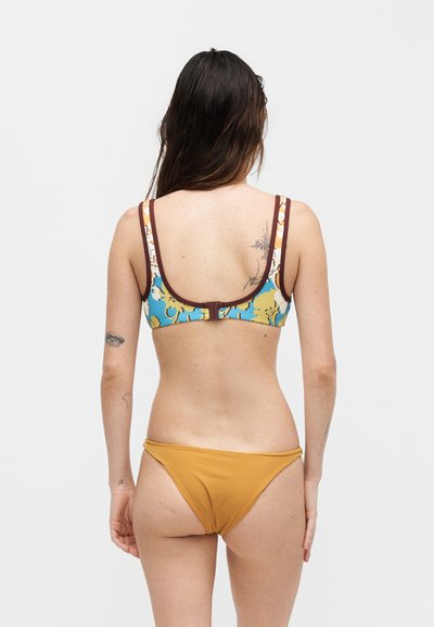 Γυναίκα με μακριά σκούρα μαλλιά δείχνει την πλάτη της, φορώντας μπλε και καφέ φλοράλ τοπ μπικίνι και μονόχρωμο μουσταρδί κάτω μέρος μπικίνι σε μονόχρωμο φόντο.