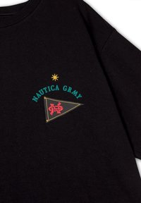 Camiseta de algodón negra con el texto "NAUTICA GRMY" en teal, una estrella amarilla y un gráfico de banderín rojo. Textura suave y corte relajado.
