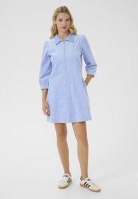Una donna sta in piedi con sicurezza, indossando un vestito azzurro chiaro con colletto e maniche a sbuffo, abbinato a scarpe da ginnastica di tendenza.