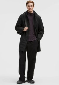 Ung mand står iført en sort frakke, mørklilla quarter-zip skjorte, sorte cargo-bukser og grå sneakers, med den ene hånd i lommen.