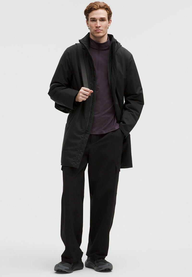 Ung mand står iført en sort frakke, mørklilla quarter-zip skjorte, sorte cargo-bukser og grå sneakers, med den ene hånd i lommen.