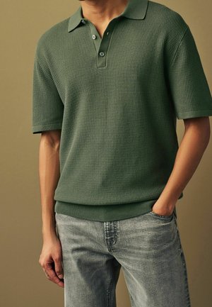 Groen gestructureerd poloshirt met een korte kraag, een twee knopen brede plooi en geribde manchetten. Draagt het met lichtgrijze spijkerbroek, handen in de zakken.