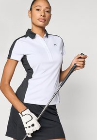 Γυναικείο πόλο γκολφ σε λευκό με μαύρες λεπτομέρειες. Ύφασμα που απομακρύνει την υγρασία, γιακάς, κοντά μανίκια, συνδυασμένο με σκουρόχρωμη φούστα και γάντι.