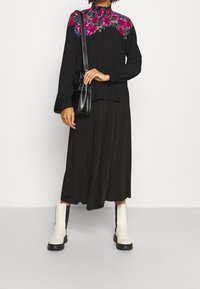 Persona che indossa una blouse nera con dettagli floreali, una gonna nera e stivaletti bianchi, mentre tiene una piccola borsa nera.