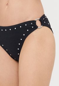 Bas de bikini noir avec des clous en argent et un anneau métallique rond accentuant le côté. Présente un tissu texturé et des détails froncés.