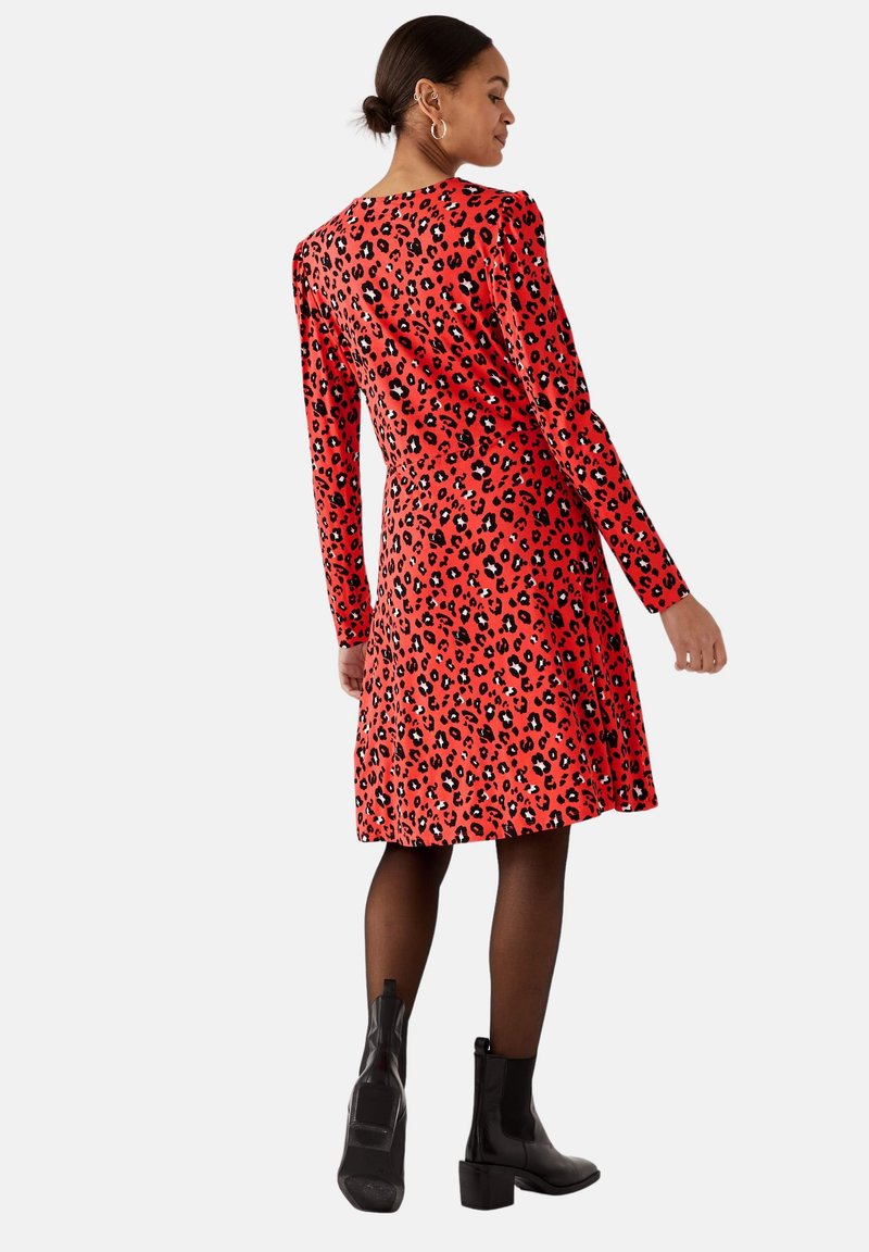 Frau in einem roten Leopardenmuster-Langarmkleid, schwarzen Strumpfhosen und schwarzen Knöchelstiefeln, die mit dem Rücken zur Kamera steht.