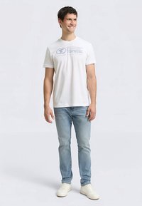 Bela bombažna majica z okroglim izrezom in potiskanim logotipom, kombinirana s svetlo modrimi kavbojkami iz jeansa in belimi supergami. Oseba stoji v nevtralnem položaju.