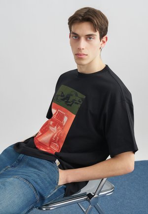 Giovane uomo con capelli castani corti indossa una t-shirt nera con grafica e jeans blu, seduto di lato su una sedia di metallo contro uno sfondo semplice.