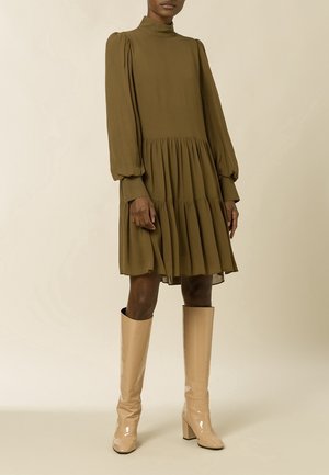 Model draagt een olijfgroene lange mouwen jurk met laagjes, gecombineerd met kniehoge glanzende beige laarzen tegen een neutrale achtergrond.