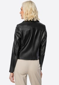 Donna con capelli biondi corti che indossa una giacca di pelle nera e pantaloni beige, di spalle su uno sfondo bianco.
