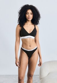 Crni pamukasti grudnjak i donji set s V-izrezom. Ima bijeli elastični pojas s "Calvin Klein" logotipom. Glatka tekstura, minimalistički stil.