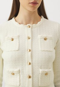 Frau trägt einen cremefarbenen strukturierten Cardigan mit goldenen Knöpfen und vier kleinen vorderen Taschen, der kurz geschnitten ist, um Hals und oberen Oberkörper zu zeigen.