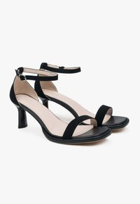 Zwarte suede hoge hakken sandaaltjes met twee banden over de tenen en een enkelbandje. Gesloten, ronde hak met een gladde, zwarte zool.