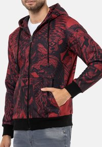 Roter Reißverschluss-Hoodie mit schwarzen gemusterten Grafiken, einfarbige schwarze Bündchen und Taillenbund, mit einer Kapuze mit Kordelzug und seitlichen Taschen.