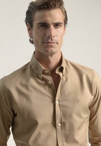 Camisa de botones marrón claro con collar, con costuras sutiles, una textura suave y un pequeño logo en el pecho.