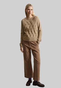 Maglione beige a trecce, con orlo a coste largo, abbinato a pantaloni marroni a vita alta e gamba dritta, e scarpe di tela scure con lacci.