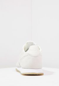 Vita mockasin och lädersneaker med gummisula, sedd bakifrån på en vit yta mot en vit hörnbakgrund.