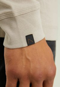 Beige gestrickter Sweatshirtärmel mit einem schwarzen Etikett und einem orangefarbenen Designstich. Glatte Textur mit geripptem Bündchendetail.