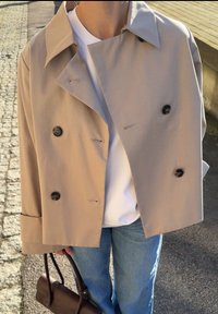 Beżowy dwurzędowy trencz, mieszanka bawełny, szerokie klapy, czarne guziki, noszony na białej koszuli, w połączeniu z jasnoniebieskimi dżinsami i brązową torebką.