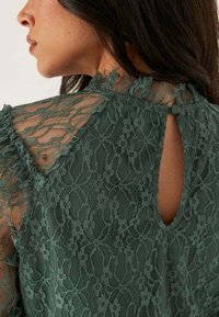 Blusa verde in pizzo con motivo floreale, colletto arricciato e chiusura con oblò sul retro con un dettaglio a bottone. Tessuto a trama, trasparente.