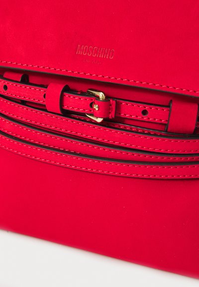 Sarkans ādas soma ar teksturētu apdari, izteiksmīgu Moschino logo un dekoratīvu jostu ar zelta sprādzi.