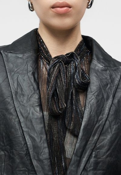 Μαύρο δερμάτινο σακάκι με υφασμένους γιακάδες· το στρωμένο τοπ έχει μαύρη και μεταλλική γραβάτα. Λαμπερό γραμμικό μοτίβο που δημιουργεί έντονη αντίθεση.