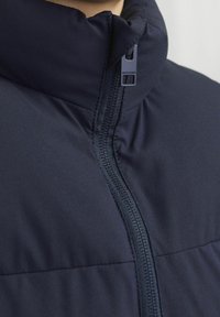 Marineblaue Steppjacke mit hohem Kragen, Frontreißverschluss und Markenreißverschlussanhänger. Glatte Textur mit dezenten gesteppten Details.