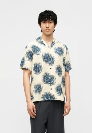 Jonge man draagt een beige overhemd met korte mouwen met blauwe zonnestraalpatronen en donkergrijze broek, staand tegen een effen achtergrond.