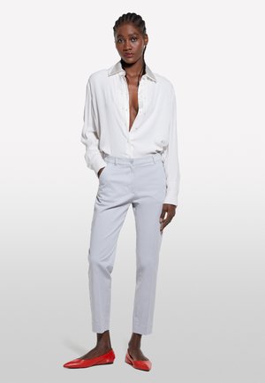 Chemise blanche à boutons, avec un décolleté en V profond, pantalon léger gris cintré, et des ballerines rouges avec des accents à œillets. Tissu lisse et coupe structurée.