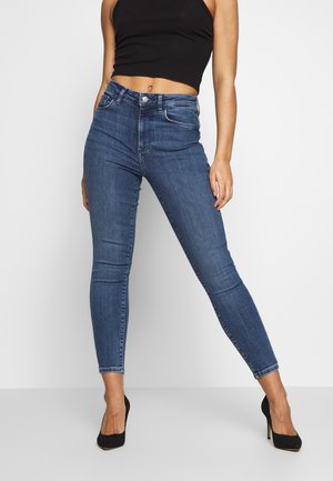 Mujer con vaqueros pitillo azules de cintura alta, top corto negro sin mangas y tacones altos negros, de pie con las piernas separadas sobre un fondo blanco.