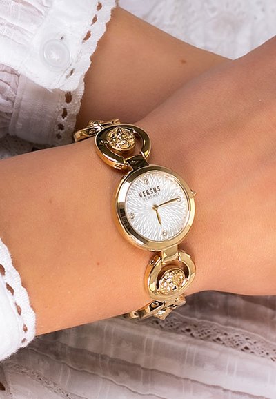 Reloj dorado con una esfera redonda, dial texturizado blanco y una pulsera decorativa enlazada con acentos circulares y florales.