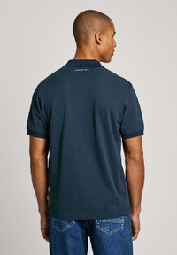 Marineblauwe polo met korte mouwen, zichtbare stiksels en een klein "LONDON W11"-opschrift op de achterkant dichtbij de kraag. Zacht katoen.