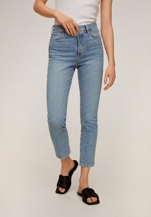 Kobieta w jasno niebieskich obcisłych dżinsach z wysokim stanem, białej bezrękawnikowej bluzce i czarnych plecionych sandałach wsuwanych, stojąca na neutralnym tle.