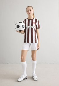 Brązowa i biała pasiasta koszulka piłkarska z logo, białe spodenki, białe skarpetki oraz czarne korki. Trzyma klasyczną czarno-białą piłkę nożną.