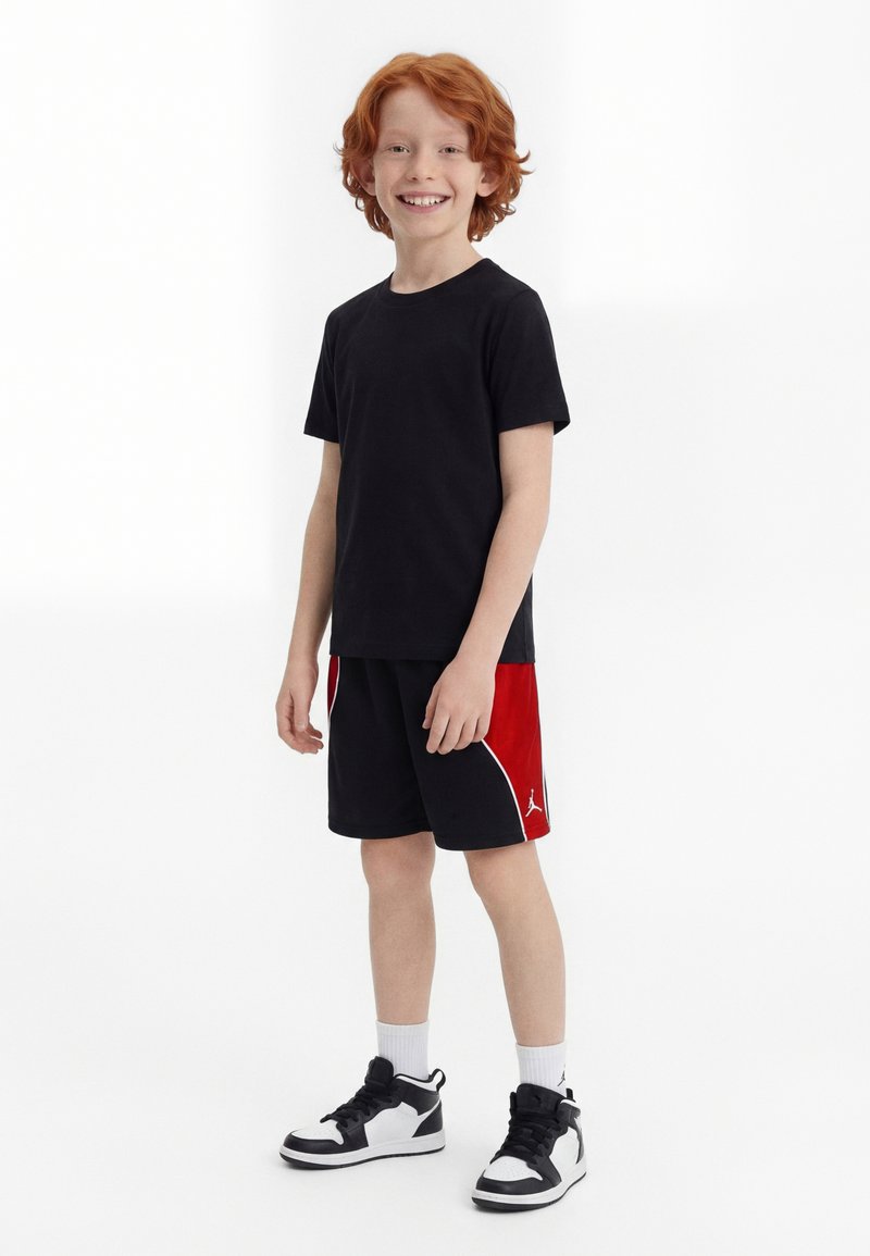 Χαμογελαστό αγόρι με κόκκινα μαλλιά που φοράει μαύρο μπλουζάκι, μαύρο και κόκκινο σορτς, λευκές κάλτσες και ασπρόμαυρα αθλητικά παπούτσια, στέκεται σε λευκό φόντο.