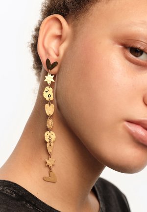 Zbliżenie osoby noszącej długą złotą wiszącą kolczyk z zawieszkami w kształcie serca, gwiazdy, twarzy, słońca i uśmiechu.