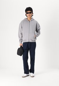 Γκρι ζακέτα με φερμουάρ και μπροστινές τσέπες, σε συνδυασμό με ναυτικές παντελόνες. Το μοντέλο κρατά μια μικρή μαύρη τσάντα και φορά μαύρα γυαλιά ηλίου. Λευκά αθλητικά παπούτσια.