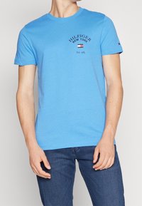 Muž oblečený v světle modrém tričku s krátkým rukávem s nápisem a logem "Hilfiger New York Est. 1985", spárován s tmavě modrými džíny.