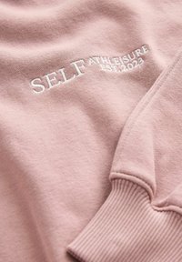Felpa rosa chiaro con polsini a coste, caratterizzata da ricami bianchi che riportano la scritta "SELF ATHLEISURE EST.2022" sul fronte. Tessuto morbido.