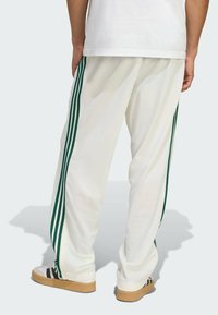 Pantalons de sport blancs avec trois bandes vertes le long des côtés, offrant une coupe décontractée et une texture douce. Des baskets beiges visibles complètent le look.