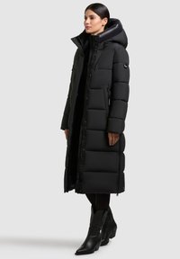 Manteau rembourré noir long avec un col haut et une capuche, présentant un design matelassé, une fermeture éclair frontale et des poches latérales.