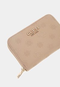 Beige portemonnee met reliëf bloemenpatronen, gouden hardware en een ritsclosure. Heeft het logo "GUESS EST 1981" prominent weergegeven.