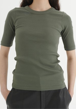Mujer con una camiseta entallada de manga corta, acanalada y de color verde oliva y pantalones negros con las manos en los bolsillos, mostrada desde el cuello hasta las caderas.
