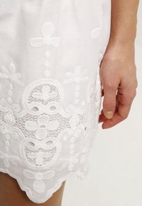 Detail eines weißen bestickten Kleides, das florale Muster, transparente Spitze, einen gewellten Saum und eine weiche Textur zeigt. Eine Hand ist am Rand sichtbar.