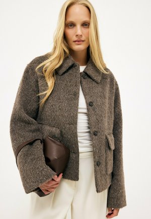 Mujer rubia con una chaqueta marrón con textura y botones sobre una camiseta blanca, sosteniendo un bolso de mano marrón oscuro.
